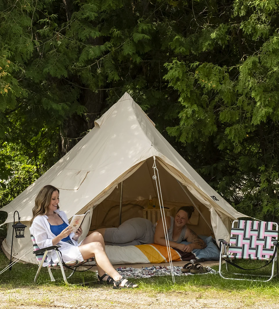 Two ladies, one in a tent and another reading a book in a chair in the woods